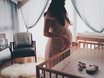 Photo by Ömürden Cengiz woman in white lace sleeveless dress standing beside brown wooden crib